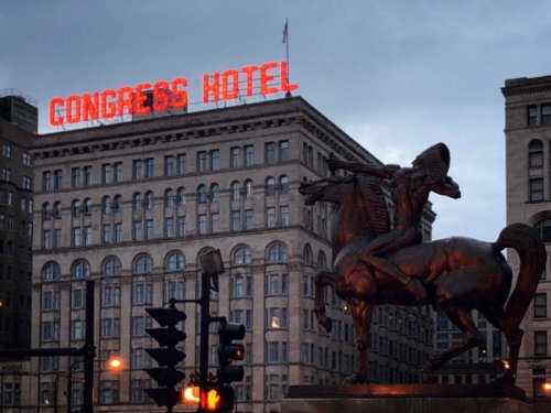 A bronze statue of a horse and rider in front of the Congress Hotel, with its neon sign illuminated at dusk.