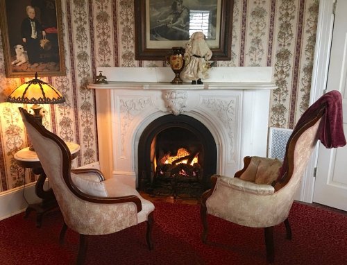 Cozy room with two armchairs facing a fireplace, vintage decor, and a lamp beside a floral wallpapered wall.