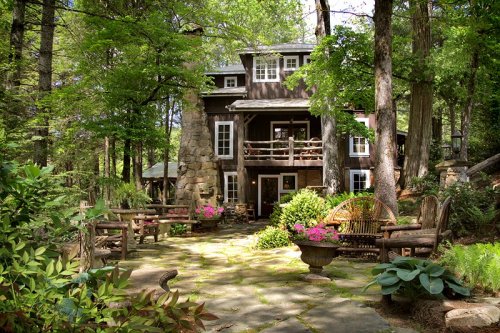 A charming rustic house surrounded by lush greenery and colorful flowers, with a stone pathway and outdoor seating.