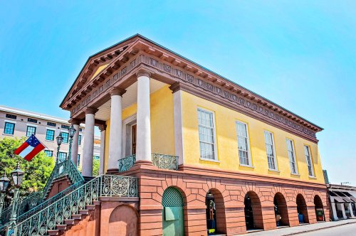 Historic building with yellow walls, white columns, and a staircase, featuring flags and a clear blue sky.