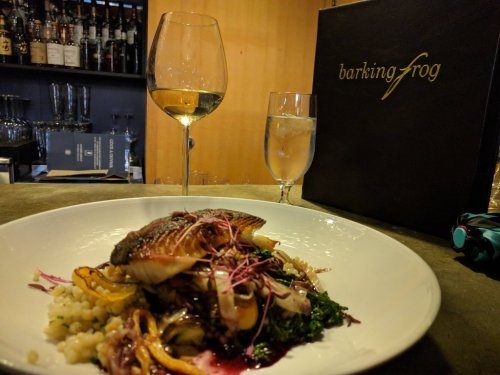 A plate of fish with vegetables and grains, accompanied by a glass of white wine and a water glass, with a menu in the background.