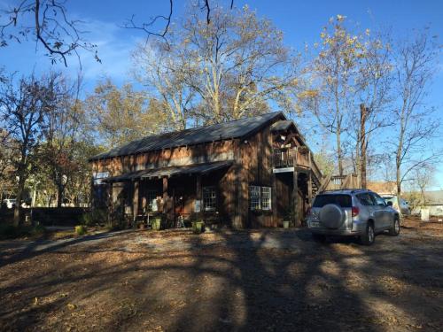 A rustic wooden house surrounded by trees, with a gravel driveway and a parked SUV in the foreground.