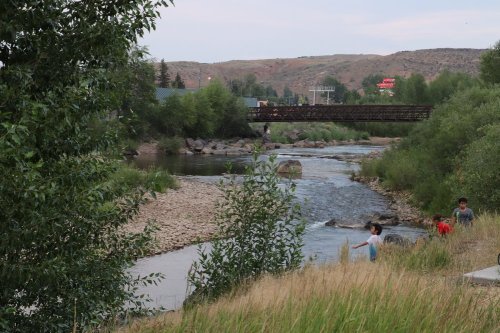 Children play by a riverbank with a bridge in the background, surrounded by greenery and gentle hills.