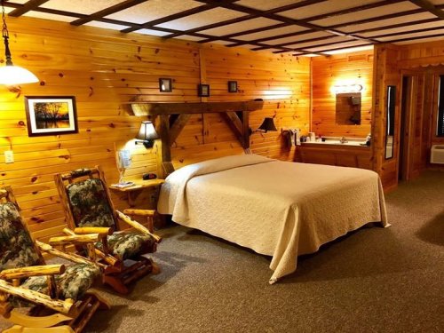 Cozy wooden cabin interior featuring a queen bed, rocking chairs, and a bathroom area with warm lighting.