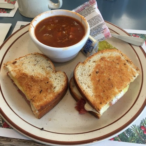 A plate with a grilled cheese sandwich, a bowl of tomato soup, and a pickle on the side.