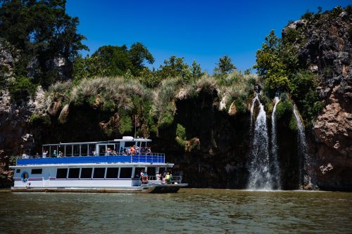 A tour boat with passengers sails past a rocky cliff with a waterfall and lush greenery under a clear blue sky.