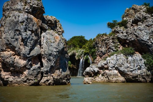 A serene landscape featuring a waterfall cascading between rocky cliffs under a clear blue sky.