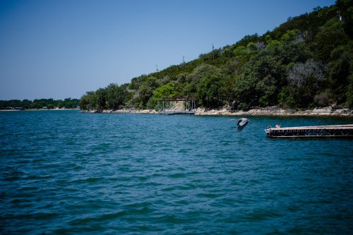 A serene lake scene with a dock, lush greenery, and a bird flying over the water under a clear blue sky.