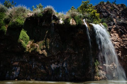 A waterfall cascading down a rocky cliff, surrounded by lush greenery under a clear blue sky.