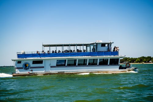 A large white boat with a blue canopy cruising on a sunny lake, with people enjoying the ride on the upper deck.