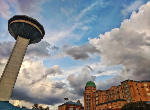 A tall water tower stands beside a multi-story building under a dramatic sky filled with clouds.