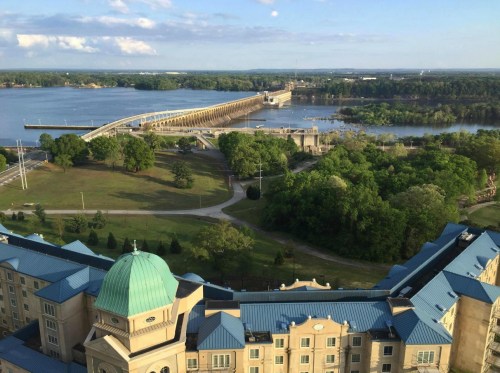 Aerial view of a river, bridge, and green parkland with a building in the foreground under a clear blue sky.