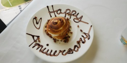 A dessert on a plate with "Happy Anniversary" written in chocolate sauce and a small cake topped with nuts.