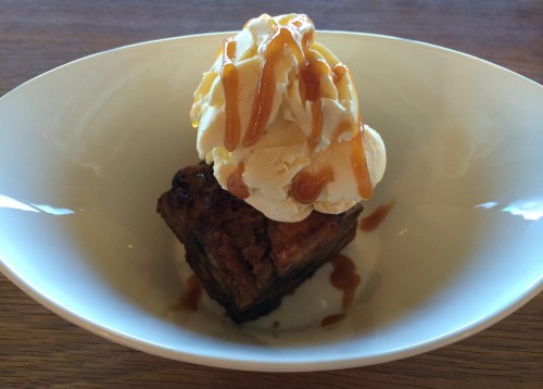 A slice of dessert topped with vanilla ice cream and drizzled with caramel sauce in a white bowl.