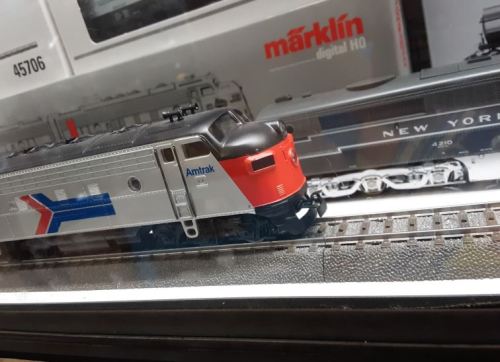A model train on tracks, featuring an Amtrak design with a red and silver color scheme, displayed in a glass case.