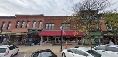 A street view of a small town with brick buildings, shops, and parked cars under a cloudy sky.