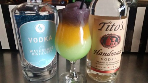 A colorful layered cocktail in a glass, flanked by bottles of Watershed and Tito's vodka on a bar counter.
