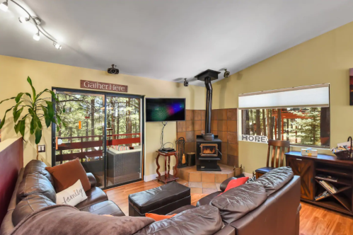 Cozy living room with a fireplace, large windows, and comfortable seating, surrounded by trees outside.