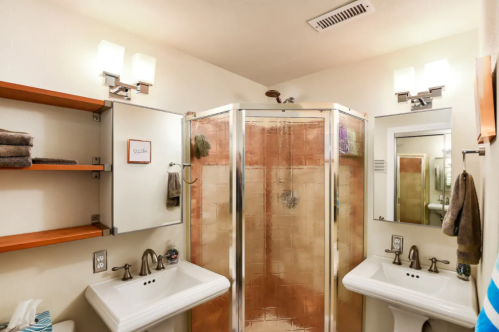 A modern bathroom featuring a glass shower, two sinks, shelves with towels, and bright lighting.