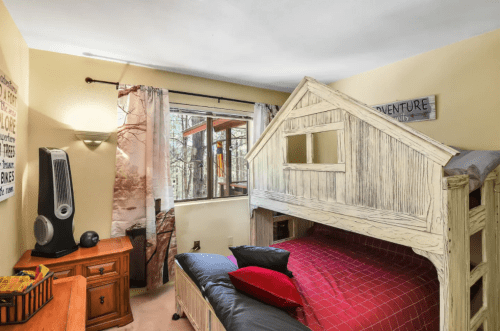 Cozy bedroom with a wooden loft bed, red bedding, and a window overlooking trees. Decor includes adventure-themed wall art.