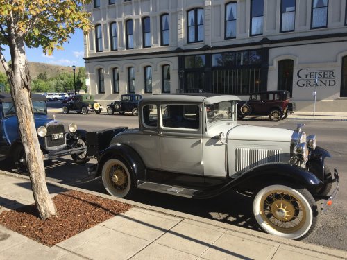 A vintage gray car parked on a street with historic buildings and other classic cars in the background.