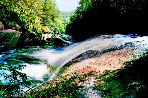 A serene river flows over rocks, surrounded by lush greenery and sunlight filtering through the trees.