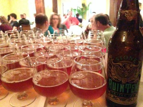 A table filled with glasses of amber beer, with a large bottle of Stone Ruination IPA in the foreground.