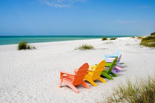 Colorful beach chairs lined up on a sandy shore, with gentle waves and clear blue skies in the background.