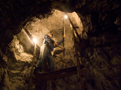 A miner using a pickaxe on a rocky surface inside a dimly lit cave, standing on a wooden ladder.