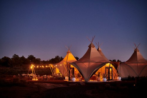 Tents illuminated at dusk, surrounded by trees, with string lights and a dining area set up outside.