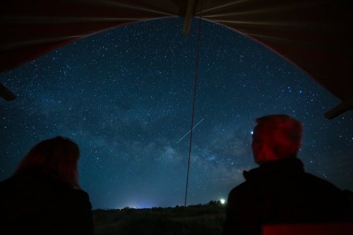 Two people sit under a shelter, gazing at a starry night sky filled with the Milky Way and a shooting star.
