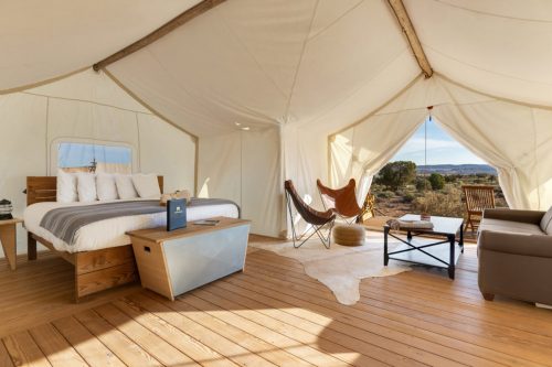 A cozy glamping tent interior with a bed, seating area, and large windows showcasing a scenic outdoor view.