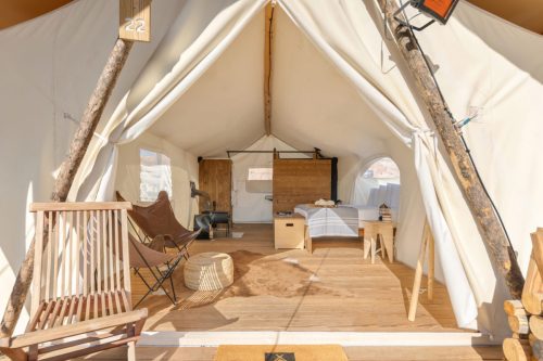 Cozy glamping tent interior with wooden furniture, a bed, and seating, surrounded by natural light.