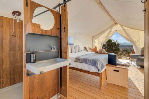 A cozy glamping tent interior featuring a bed, wooden accents, and a small sink area with a mirror. Natural light floods in.