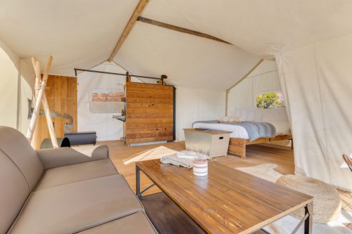 Cozy glamping tent interior with a bed, sofa, wooden table, and rustic decor. Bright and inviting atmosphere.