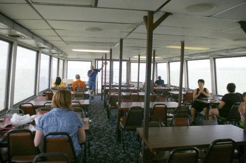 Interior of a dining area on a boat, with tables, chairs, and large windows overlooking the water. People are seated and dining.