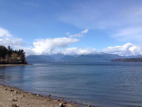 A serene lake scene with mountains in the background, under a blue sky with fluffy clouds and a pebbly shoreline.