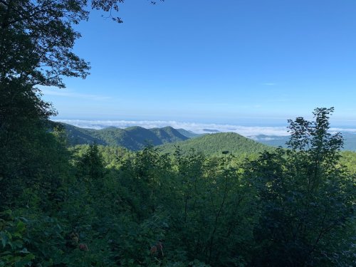 A scenic view of green mountains under a clear blue sky, with clouds hovering in the valleys below.