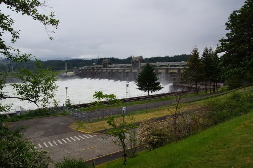 A large dam releasing water, surrounded by trees and a cloudy sky, with a road and parking area in the foreground.
