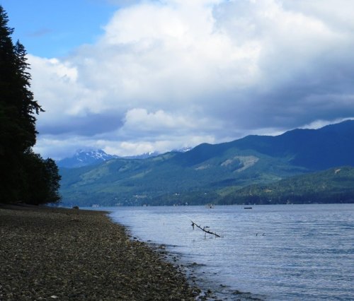 A serene lakeside view with mountains in the background, under a partly cloudy sky. Pebbly shore and calm water.