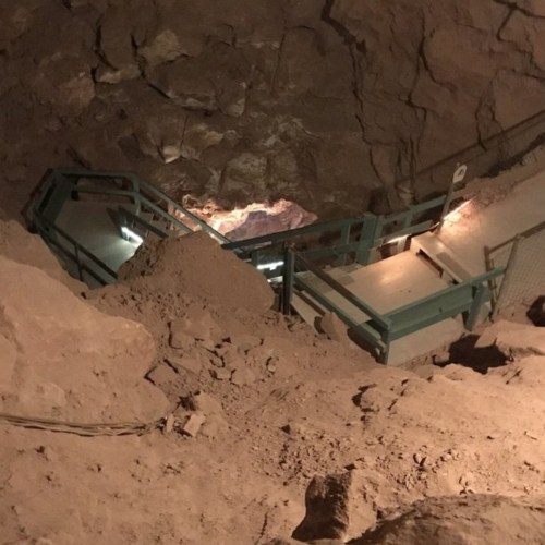 A dimly lit cave with a staircase and a sloped path, surrounded by rocky walls and loose debris.