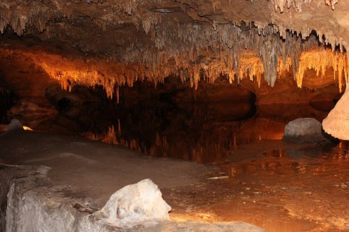 A dimly lit cave with stalactites hanging from the ceiling and a reflective pool of water on the ground.