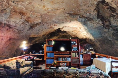 Cozy cave interior with a sofa, bookshelves, a TV, and a dining table, illuminated by warm lighting.