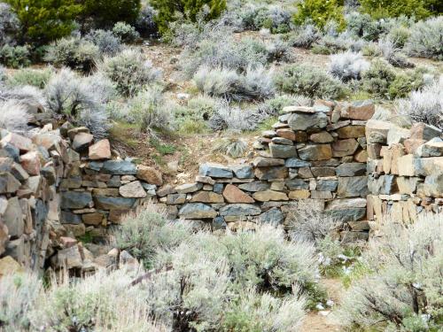 Ruins of a stone wall surrounded by sagebrush and greenery in a natural landscape.