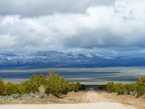 A dirt road leads to a vast landscape with mountains in the distance, under a cloudy sky.