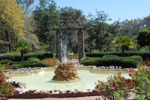 A serene garden featuring a stone fountain surrounded by lush greenery and colorful flowers under a clear blue sky.