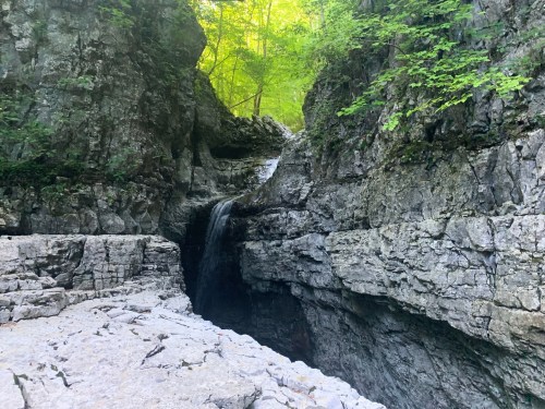 A serene waterfall cascades between rocky cliffs, surrounded by lush green foliage in a tranquil natural setting.