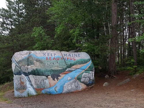 A large painted rock in a forest, featuring the words "KEEP MAINE BEAUTIFUL" and a scenic landscape with a bear.