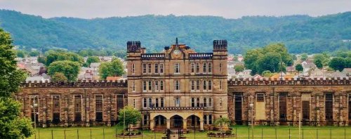 Historic stone building with towers, surrounded by greenery and mountains in the background.