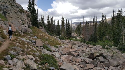 A person walks along a rocky trail surrounded by tall trees and mountains under a cloudy sky.